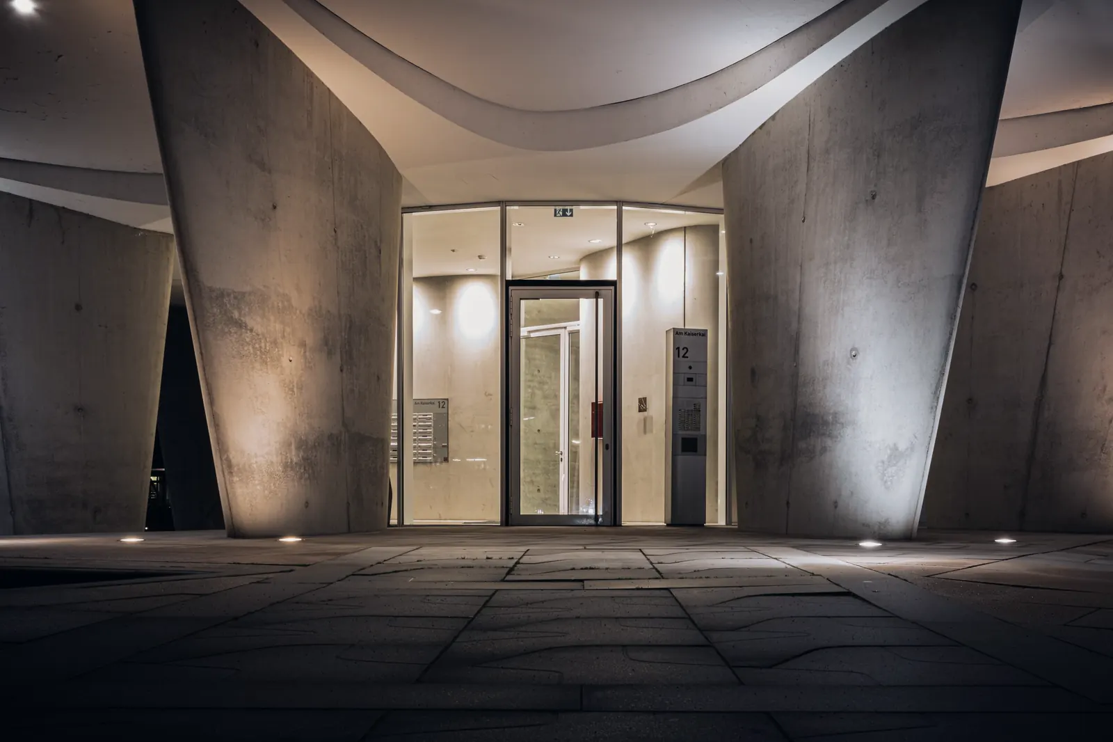 Entrada entre dos muros curvos de hormigón con una puerta de vidrio al fondo.